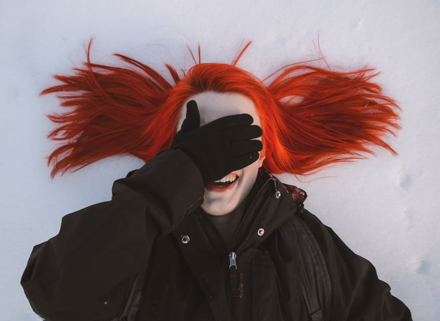 Joyful redhead woman in winter clothing laughing playfully while covering her face with a gloved hand on a snowy background.