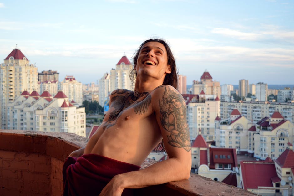 Smiling tattooed man on a balcony enjoying a cityscape view under a clear sky.