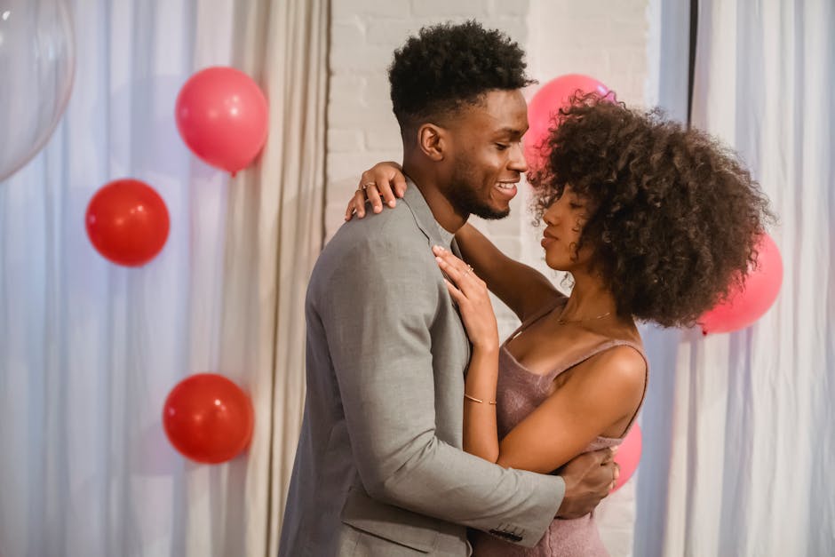 Side view of young black couple in elegant clothes standing in bright room and cuddling while dancing together with closed eyes near pink balloons on wall and curtains