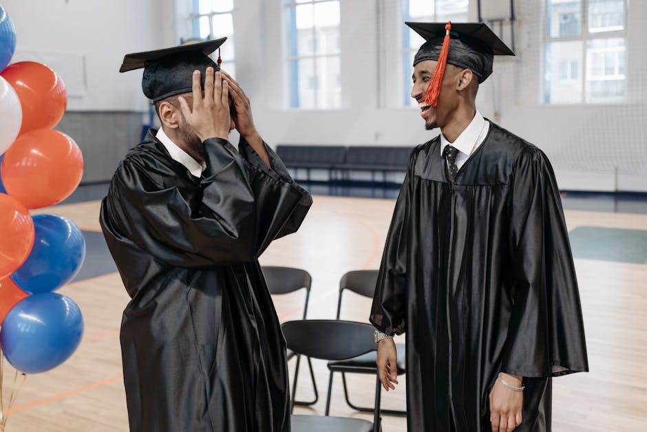 Two graduates sharing a joyful moment in caps and gowns indoors.