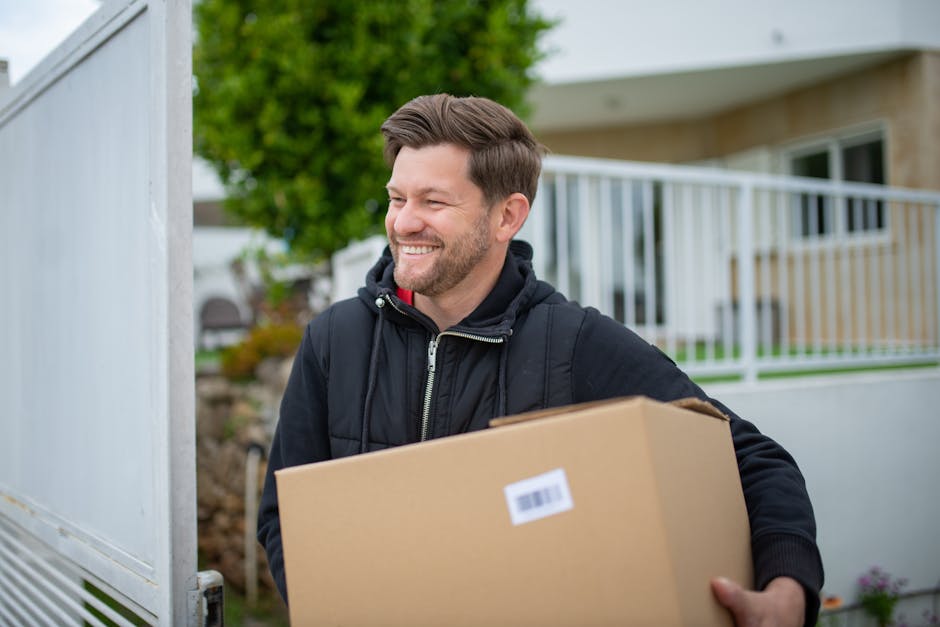 A man in a black jacket smiles while carrying a cardboard box outside a house.