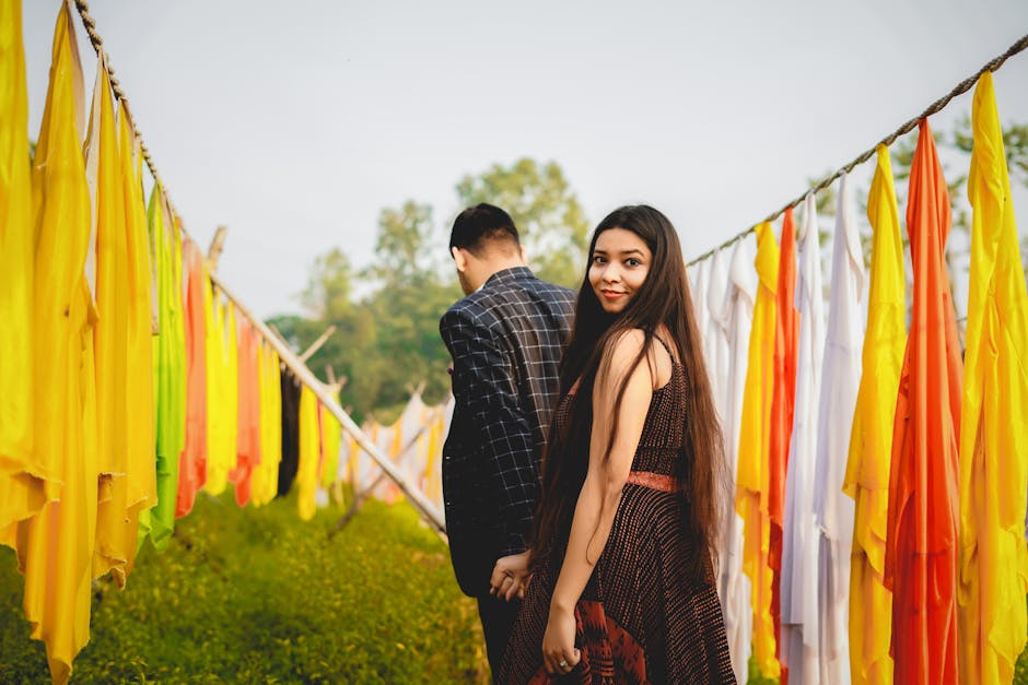A young couple holding hands, walking through colorful cloths in a serene outdoor setting.