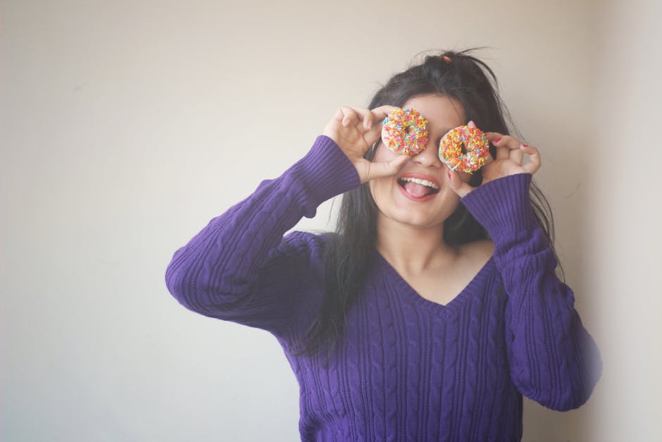 Smiling woman holding colorful donuts playfully over her eyes indoors.