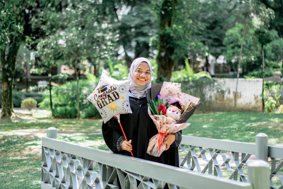 A happy graduate in hijab holding flowers and balloons in a scenic park.