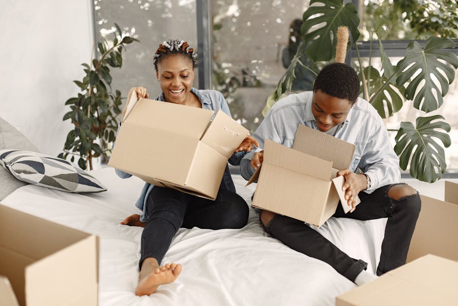 A joyful couple sits on a bed, opening boxes during a home move.
