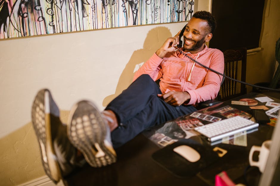 Smiling man in casual attire working from home, talking on the phone with feet up on the desk.