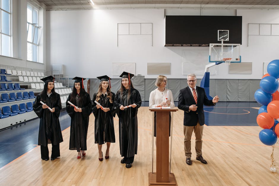 Young graduates in gowns receiving diplomas during a ceremony in a gymnasium setting.