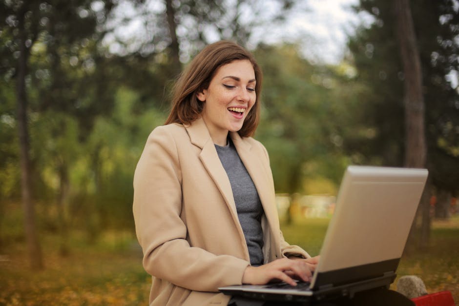 Smiling woman using laptop for remote work in a scenic autumn park setting.