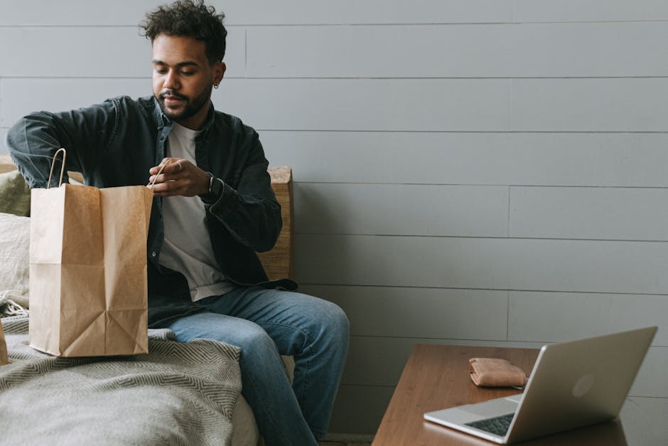 Young man unpacking delivery bag in modern home setting, seated near laptop.