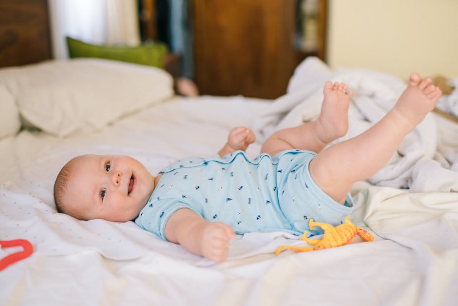 Adorable baby in blue onesie lying on bed with toy dinosaur, smiling brightly.