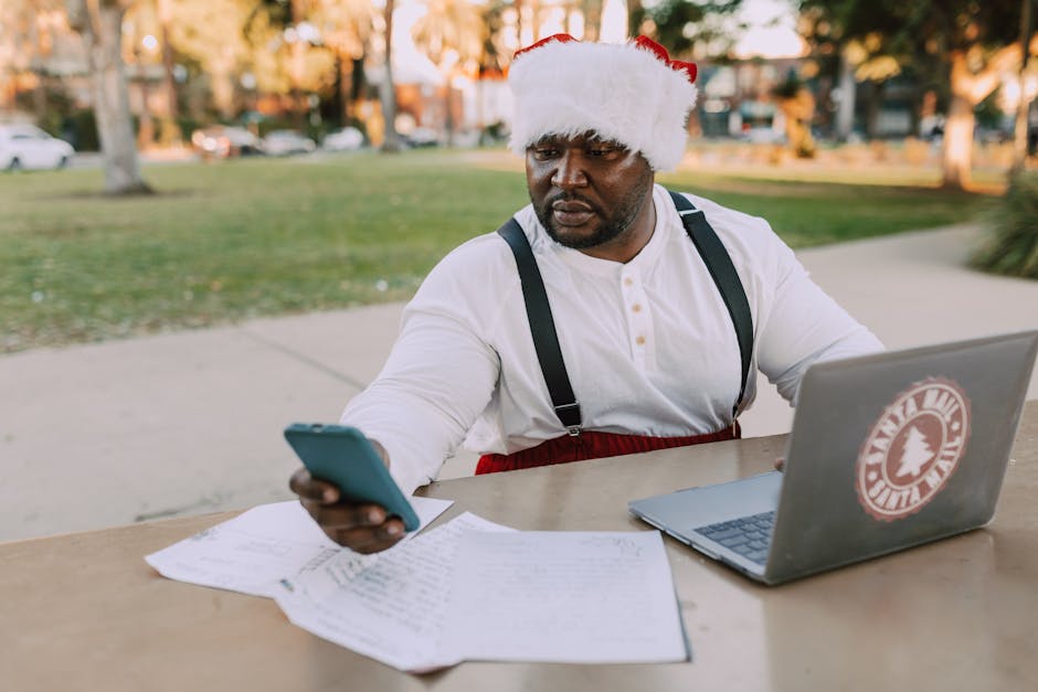 Man wearing a Santa hat using a smartphone and laptop outdoors in a festive setting.