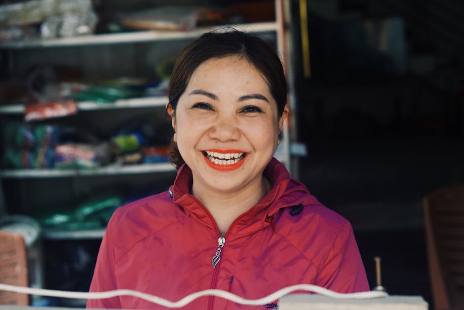 A cheerful woman in a pink jacket smiling broadly, standing in front of a market stall.