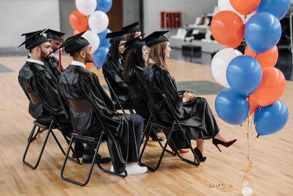 A group of graduates in caps and gowns seated indoors with colorful balloons nearby.