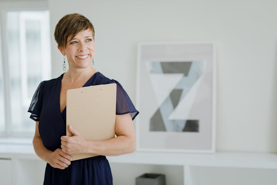 Caucasian woman smiling while holding a clipboard in an office setting, wearing a blue dress.