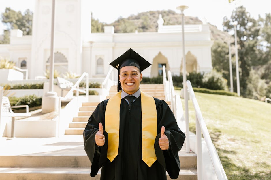 Young graduate in cap and gown giving thumbs up, celebrating success outdoors.