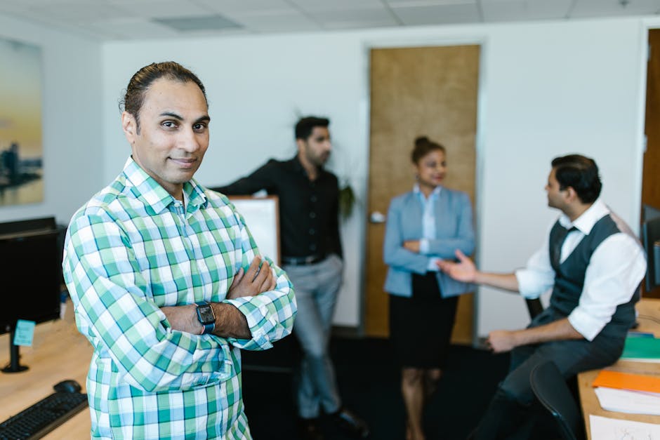 Smiling businessman with crossed arms in an office, with colleagues discussing in the background.