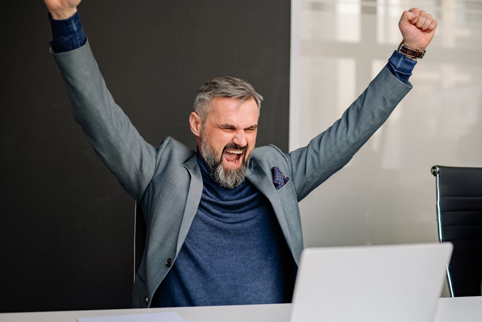 Thrilled businessman with raised arms celebrating success at his office desk.