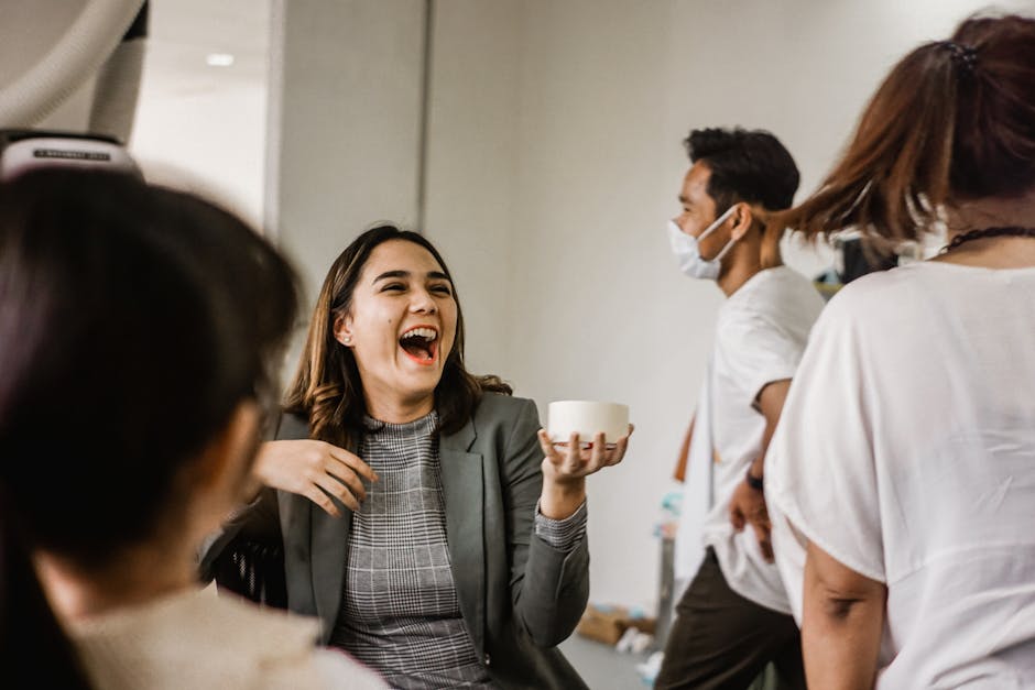A woman in a gray blazer shares a laugh with colleagues in a lively office setting.