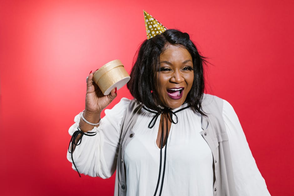 Happy woman in party hat holding a gift box with a big smile against a red background.