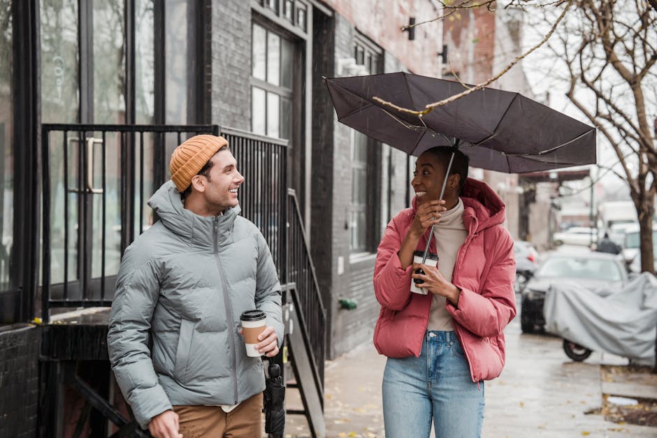 Positive African American woman with umbrella and man looking at each other while standing with takeaway coffee on rainy and windy day