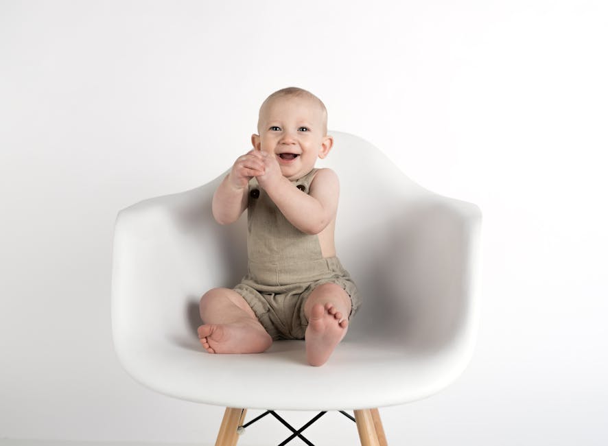 Adorable baby boy in beige romper joyfully sitting on a modern white chair indoors.