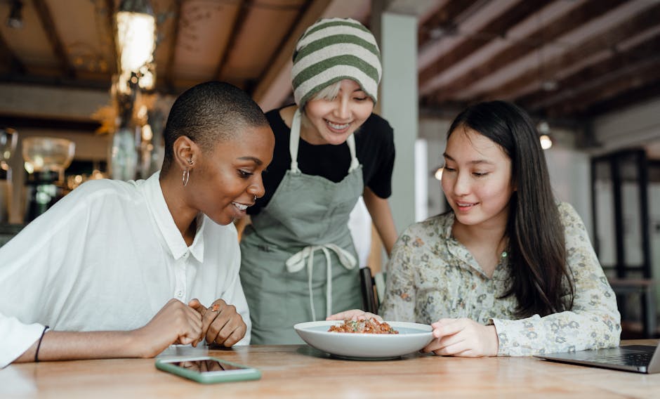 Smiling positive multiracial females in casual clothes sitting at table in modern cafe near cheerful waitress and looking at delicious dish
