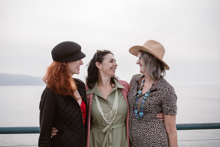 Three women in stylish outfits smiling together outdoors by the sea.