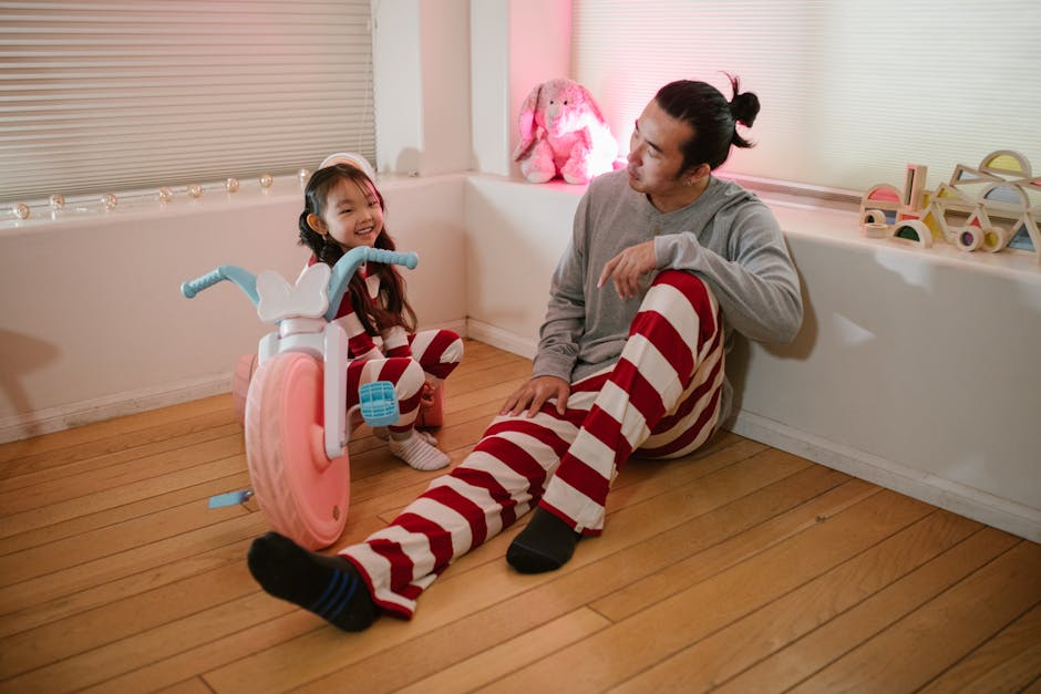 A father and daughter in matching striped pajamas share a playful moment indoors with a toy bike.