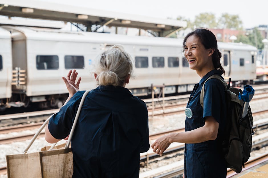 Two women laughing and talking outdoors at a busy train station during the day.