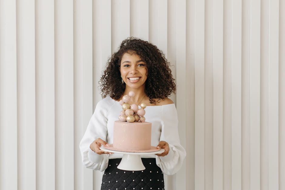 A cheerful woman with curly hair holding a cake and smiling indoors against a modern backdrop.
