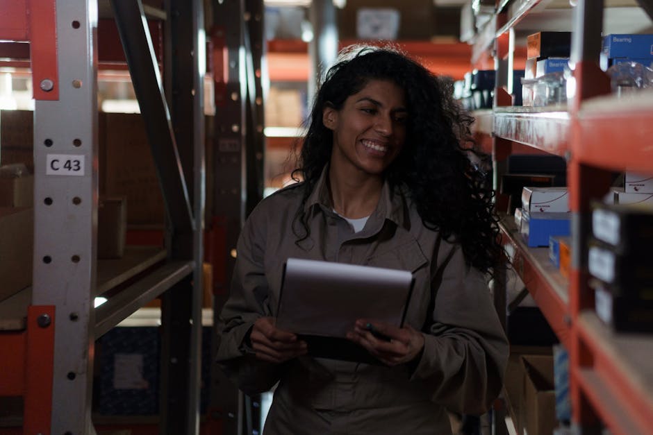 Woman managing inventory with clipboard, smiling in a dimly lit warehouse aisle.